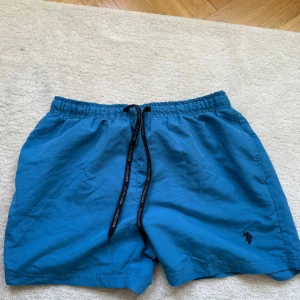 Blå badbyxor från U.S. Polo Assn. - Säljer ett par blå badbyxor från U.S. Polo Assn. med elastisk midja och snörning. De har en liten broderad logga på framsidan och en bakficka. Perfekta för sommarens alla äventyr!