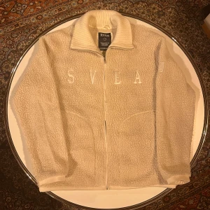 Beige fleecejacka från Svea - Mysig beige fleecejacka från Svea med dragkedja och broderad text på framsidan. Jackan har en hög krage och två fickor framtill. 
