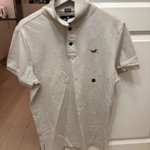 Ny beigegrå pikétröja från Hollister, med etikett kvar  - Säljer en helt ny  grå pikétröja från Hollister med korta ärmar och knappdetaljer vid kragen. Tröjan har en liten broderad logga på bröstet. Butikspris ~300:- 