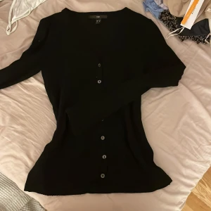 Svart kofta från H&M - Säljer en stilren svart kofta från H&M i storlek M. Koftan har långa ärmar och knappar framtill, perfekt för lager-på-lager-stil. Passar till många olika outfits och tillfällen.
