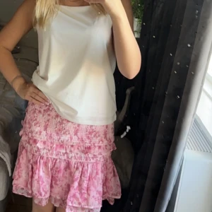 Rosa volangkjol med blommigt mönster - Supersöt rosa kjol med volanger och blommigt mönster. Kjolen är kort och har en smockad midja som ger en bekväm passform. Perfekt för dig som gillar en feminin och somrig stil.  passar mig som brukar ha s-m men är stretchig 