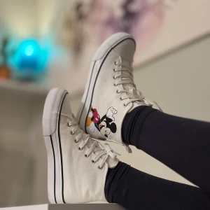 Vita Disney sneakers med Mimmi och Musse Pigg - Säljer ett par vita höga sneakers med tryck av Mimmi och Musse Pigg på sidorna. Skorna har snörning, svart rand längs sulan och är tillverkade i 100% bomullscanvas. Perfekta för dig som älskar Disney och vill sticka ut med din stil!