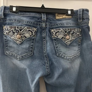 Blå jeans från Miss Me med broderade bakfickor - Säljer ett par blå jeans från Miss Me med snygga broderade detaljer och knappar på bakfickorna. Jeansen har en klassisk passform och är tillverkade i denim med coola slitningar och kontrastsömmar. Det finns lite slitage på byxorna men inte så illa, byxorna är uppsprättade längst ner. Mått innebenslängden 72cm 