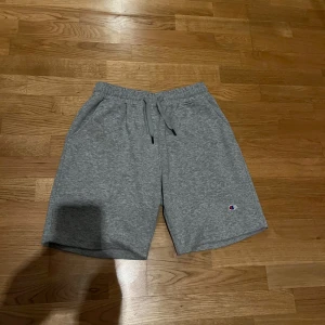 Grå shorts från Champion - Gråa shorts från Champion med elastisk midja och dragsko. Klassisk logga broderad på vänster ben. Mjukt material och två sidofickor, perfekt för en avslappnad stil.