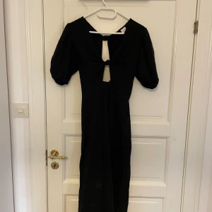 Svart långklänning med puffärmar från H&M - Säljer en svart långklänning från H&M med puffiga kortärmar och snygg knytning framtill. Klänningen har en öppen detalj vid bröstet och är tillverkad i ett mjukt material. Perfekt för dig som vill ha en stilren och trendig look.