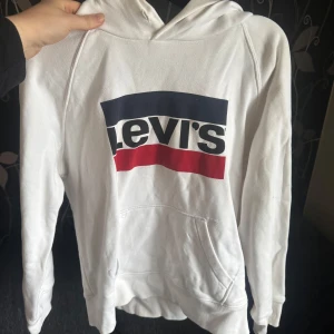 Vit hoodie från Levi's - Säljer en vit hoodie från Levi's med stor logga i blått, vitt och rött framtill. Tröjan har huva och en stor magficka. Perfekt för en avslappnad och stilren look.