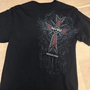 Vintage Hard Rock Cafe  - Cool svart t-shirt från Hard Rock Cafe Bengaluru med stort tryck av kors, vingar och rustning både fram och bak. Köpt på en vintage marknad i nyskick. Tillverkad i 100% bomull och har en baggy passform. Perfekt för dig som gillar rockiga emo motiv och unika t-shirts.
