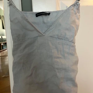 Ljusblå linnetopp från Brandy Melville - Supersöt ljusblå linnetopp från Brandy Melville med smala axelband och v-ringning. Toppen har knyt i ryggen för en justerbar passform och är tillverkad i ett tunt, svalt material. Perfekt för varma dagar och enkel att styla med jeans eller kjol.