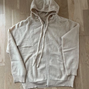 Beige stickad hoodie med dragkedja - Mysig beige hoodie med dragkedja framtill och huva med långa snören. Tröjan har långa ärmar, ribbade muddar och två fickor framtill. Perfekt för lager-på-lager och chill dagar.