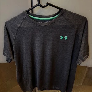 Mörkgrå t-shirt från Under Armour - Säljer en mörkgrå t-shirt från Under Armour med turkos logga på bröstet. T-shirten har korta ärmar och är i ett syntetmaterial som andas, perfekt för träning eller sport. Modellen är loose fit för extra rörelsefrihet.