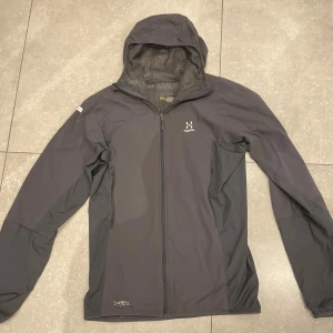 Grå vår/sommar jacka från Haglöfs. Storlek S/M - Säljer en grå vindjacka från Haglöfs med huva och hel dragkedja. Jackan har meshfoder på insidan och två sidofickor med dragkedja. Perfekt för sommaren och våren. Nypris var 1600kr. Storleken är S men passade mig även när jag hade M i storlek. Inga större defekter. Har vårdats bra. 