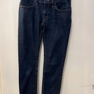 Mörkblå jeansbyxor- Tiger Of Sweden  - Säljer ett par klassiska mörkblå jeansbyxor med fem fickor och smal passform. Byxorna har normal midja och raka ben, perfekta till vardags. Tidlös design med guldfärgade sömmar och knappstängning framtill.