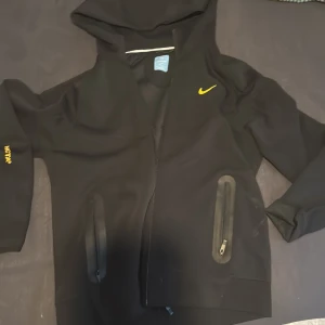 Svart hoodie från Nike NOCTA - Svart hoodie från Nike NOCTA med gul logga på bröstet och NOCTA-tryck på ärmen. Jackan har dragkedja framtill, två fickor med dragkedja och huva. Perfekt för en sportig och stilren look.
