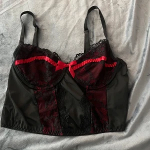 Svart och röd korsett med spetsdetaljer - Elegant svart och röd korsett med spetsdetaljer och en söt rosett framtill. Den har justerbara axelband och genomskinlig rygg har inga bilder hur den ser på då den är för liten 