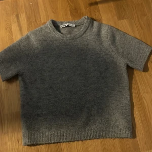 Grå stickad kortärmad tröja från Zara - Mysig grå stickad tröja från Zara med korta ärmar och rund halsringning. Perfekt för lager-på-lager och enkel att matcha till olika outfits. Ribbstickade kanter vid ärmslut och nederkant. Knappt använd och så skönt material