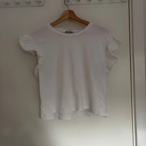 Vit topp med volangärmar - Säljer en söt vit topp med korta volangärmar. Perfekt till sommaren och enkel att matcha med olika outfits. Toppens material känns mjukt och luftigt, vilket gör den skön att bära varma dagar.
