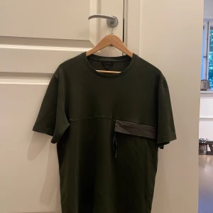 Mörkgrön t-shirt med fickdetalj från COS - Säljer en mörkgrön t-shirt från COS med en unik fickdetalj i mörkare tyg på bröstet. T-shirten har rund halsringning och korta ärmar. Perfekt för dig som gillar minimalistisk stil med en twist.