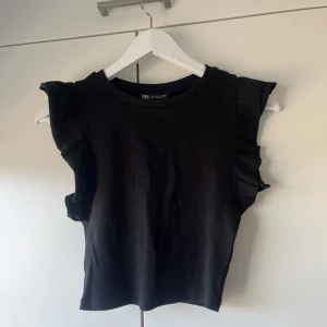 Svart topp från Zara - Snygg svart topp från Zara med volangdetaljer på axlarna. Toppen är ribbad och har en rund halsringning. 