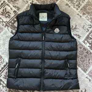 Svart dunväst från Moncler - Säljer en svart dunväst från Moncler med hög krage och dragkedja framtill. Västen har två fickor med dragkedja och Moncler-logga på bröstet. Perfekt för lager-på-lager och stilren look.
