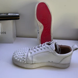 Vita sneakers med nitar från Christian Louboutin - Snygga vita sneakers från Christian Louboutin med coola nitar på tån. Skorna har snörning och en klassisk rund tå. Perfekta för att ge din outfit en edgy touch. Skick 6/10, det går att fixa till skorna, Storlek 42, allt og kommer med 