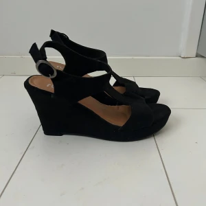 Svarta kilklackar/högklackade skor - Fina högklackade skor. Skorna har öppen tå, breda remmar och spänne vid ankeln. Perfekta för sommaren och ger höjd. Snygg och enkel design som passar till många outfits. Endast använda vid ett tillfälle. Superfint skick!