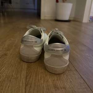Vita sneakers från Golden Goose - Säljer ett par stilrena vita sneakers från Golden Goose med grå snörning och silverdetaljer på hälen. Perfekta för en avslappnad look med en touch av lyx.