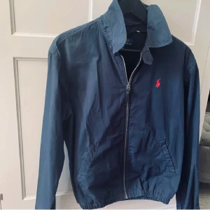 Blå vindjacka från Polo Ralph Lauren - Snygg blå vindjacka från Polo Ralph Lauren med dragkedja framtill och klassisk krage. Jackan har elastisk nederkant och är prydd med den ikoniska röda loggan på bröstet. Perfekt för en stilren och avslappnad look. Jackan är i nyskick knappt använd, säljer den för att den inte riktigt var min stil. Kom med frågor eller funderingar så är det bara o skriva ☺️ kan gå ner i pris