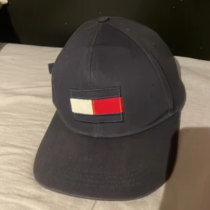 Mörkblå keps från Tommy Hilfiger - Snygg mörkblå keps från Tommy Hilfiger med klassisk broderad logga framtill i vitt, rött och blått. Justerbar rem baktill för perfekt passform. Perfekt accessoar för en sportig och stilren look.