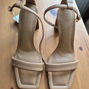 Beige klackskor från Stradivarius - Säljer ett par stilrena beige sandaler från Stradivarius med fyrkantig tå och bred klack. Helt nya, aldrig använda. Skorna har justerbart spänne runt ankeln och en minimalistisk design. Perfekta för dig som gillar en enkel men trendig look. 