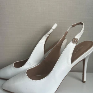 Klackar storlek 39 - Säljer ett par eleganta vita pumps från Anna Field med spetsig tå och hög klack. Skorna har öppen häl och en rem med glittrig smyckesdetalj på sidan. Perfekta för speciella tillfällen.
