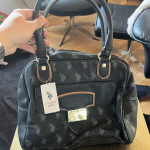 Svart handväska från U.S. Polo Assn. - Säljer en svart handväska från U.S. Polo Assn. med diskret logomönster och guldfärgat spänne framtill. Väskan har dubbla handtag, en yttre ficka med lock och detaljer i beige. Perfekt för dig som vill ha en stilren accessoar med klassisk känsla. Morsan köpte i usa med som ni ser så har den aldrig ens blivit använd och prislappen sitter kvar. $69 motsvarar ungefär 800kr :)