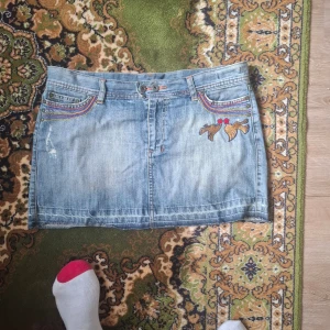 Jeanskjol med broderi och fickor - Säljer en ljusblå jeanskjol med broderade detaljer i gult och rött samt dekorativa fickor bak. Kjolen har klassisk jeanslook med knapp och dragkedja framtill och broderade motiv på ena sidan fram.