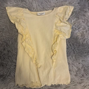 Gul ribbad topp med volang från Kappahl - Söt ljusgul ribbad topp från Kappahl med volangdetaljer längs axlarna och framtill. Toppen har korta volangärmar och är tillverkad i mjukt material, perfekt för vår och sommar.