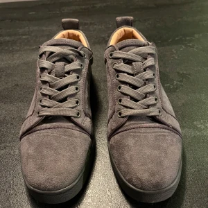 Grå mockasneakers med röd sula från Christian Louboutin - Snygga grå sneakers i mocka med klassisk röd sula från Christian Louboutin. Skorna har rund tå, platt sula och grå snörning. Insidan är ljusbeige och de har en stilren, minimalistisk design som passar perfekt till streetwear.