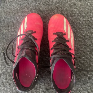 Rosa och svarta speedportal 3 fotbollsskor från Adidas - Säljer ett par rosa och svarta fotbollsskor från Adidas med vita ränder på sidorna. Skorna har snörning och dobbar under sulan för bra grepp på planen. Perfekta för dig som vill sticka ut på fotbollsplanen! Skorna är lite smutsiga men det går att få bort.
