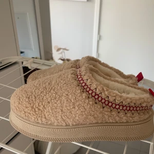 Beige fluffiga tofflor med röd detaljsöm💛 - Mysiga beige tofflor med fluffigt foder. Är gjorda för att ha ute men funkar också som inneskor! Utsidan är i mjuk teddy-liknande syntet och kanten har en röd sömm. Aldrig använda så i nyskick!