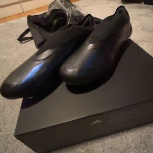 Svarta fotbollsskor från Adidas - Säljer ett par svarta Adidas fotbollsskor med en modern, strömlinjeformad design. Skorna har en slät ovandel och platt sula med dobbar, perfekta för spel på gräsplan. Snygga detaljer i mörkblått ger en extra touch.