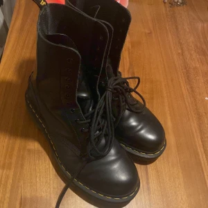 Svarta boots från Dr. Martens - Klassiska svarta Dr. Martens boots med högt skaft och gul söm runt sulan. Skorna har snörning framtill och är tillverkade i skinn med robust, mönstrad gummisula. Perfekta för dig som gillar en tidlös och edgy stil.
