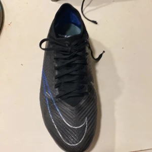 Nike fotbollsskor i svart och blå - Säljer ett par Nike PRO fotbollsskor i svart med blå och vita detaljer i storlek 42,5. Skorna har använts tidigare i 1 månad men blev tyvärr för små