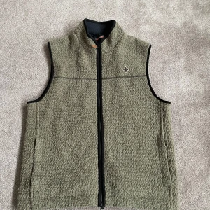 Olivgrön fleeceväst med dragkedja - Säljer en olivgrön fleeceväst med hel dragkedja framtill och hög krage. Västen har ett diskret emblem på bröstet och svarta kantband vid ärmhålen. Perfekt för lager-på-lager under kyliga dagar.