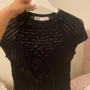 Svart stickad topp från Zara - Snygg svart stickad topp från Zara med korta ärmar och ett genombrutet mönster framtill. Toppen har rund halsringning och är figurnära i modellen. Perfekt att styla till olika outfits!