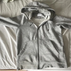 Grå hoodie med dragkedja från Jack & Jones - Säljer en klassisk grå hoodie från Jack & Jones med dragkedja framtill och två stora fickor. Tröjan har huva med snörning och är perfekt för en avslappnad stil. Passar både till jeans och träningsbyxor.