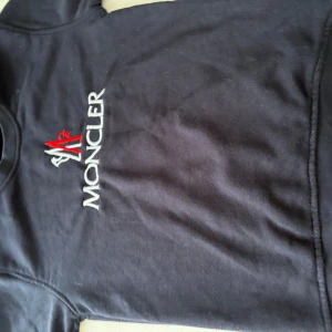 Svart långärmad t-shirt från Moncler - Svart långärmad t-shirt från Moncler med broderad logga i vitt och rött på bröstet. Klassisk rund halsringning och mjukt material som känns skönt mot huden. Perfekt för en stilren och avslappnad look.