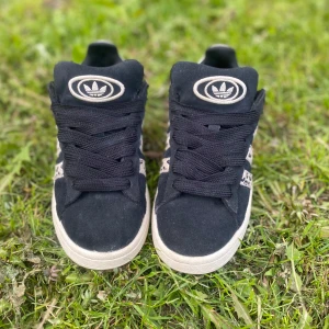 Svarta Adidas campus med leopardmönster - Säljer ett par svarta Adidas campus med vita sulor och leopardmönstrade ränder på sidorna. Skorna har svart snörning, vita snören kan medfölja vid önskemål. Perfekta för dig som vill sticka ut med en cool detalj!