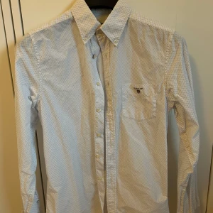 Gant skjorta - Klassisk vit skjorta från Gant med små blå prickar och button-down krage. Bröstficka med broderad logga och knäppning framtill. Perfekt för en stilren look.