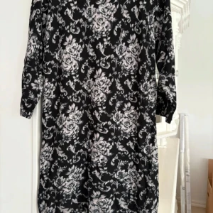 Svartvit blommig klänning från Flash Woman - Svart klänning med vitt blommönster från Flash Woman. Klänningen har lång ärm och rak passform, perfekt för dig som gillar en klassisk och stilren look med lite extra mönster.