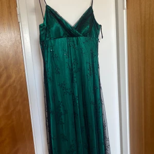 Grön långklänning med spetsdetaljer - Säljer en smaragdgrön långklänning med tunna axelband och v-ringad urringning, köpt på Humana. Klänningen har ett lager av svart spets med blommigt mönster och subtila paljetter som ger extra glans. Perfekt för balsäsong! Passar som M💚