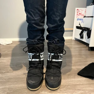 Svarta Moon Boots med vit logga - Säljer ett par klassiska svarta Moon Boots med vit logga och snörning framtill. Skorna har en rund tå, platt sula och är fodrade med vitt tyg på insidan. Perfekta för vintern och håller fötterna varma. Storlek 35/38 - jag har storlek 38
