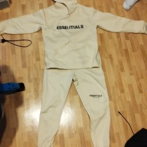 Beige Essentials hoodie från Fear of God - Säljer en beige hoodie från Essentials Fear of God med svart logga på bröstet. Hoodien har dragsko i huvan och är i mjukt material, perfekt för en avslappnad stil.
