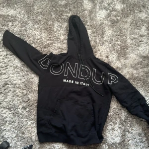 Svart hoodie från Dondup - Säljer en svart hoodie från Dondup med stor vit logga framtill och texten 'Made in Italy'. Tröjan har huva med dragsko och en stor magficka. Perfekt för en avslappnad och stilren look.
